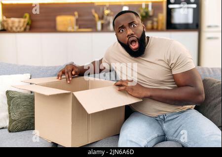 Stupito eccitato scioccato africano americano ragazzo scompatta il suo pacchetto atteso da tempo, si siede a casa sul divano in salotto, guarda la macchina fotografica in sorpresa, si sente sconcertante. Concetto di shopping online Foto Stock