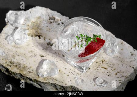 Vodka in bicchierino di ghiaccio, decorata con fette di fragola e timo sul podio in travertino su sfondo di pietra scura. Cocktail alcolico shot con stra Foto Stock