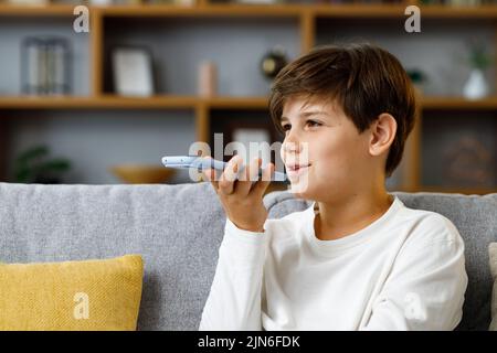 Giovane ragazzo carino che registra un messaggio vocale sullo smartphone. Sorridente adolescente che utilizza l'app Virtual Assistant seduto a casa in pullman. Riconoscimento vocale, audio Foto Stock