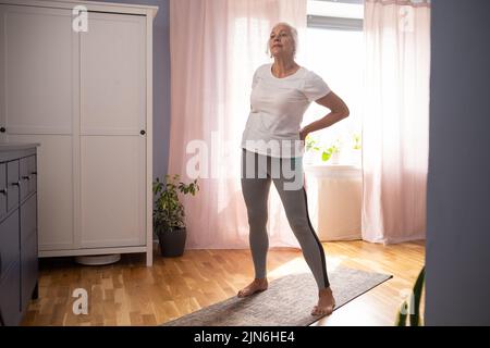 Signora anziana che fa asana yoga nel soggiorno. Foto Stock