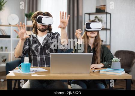 La giovane coppia si siede al tavolo, utilizzando cuffie per realtà virtuale con un moderno notebook per un'esperienza di realtà virtuale. Famiglia felice che trascorre il tempo libero a casa e gioca. Foto Stock