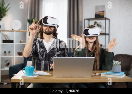 La giovane coppia si siede al tavolo, utilizzando cuffie per realtà virtuale con un moderno notebook per un'esperienza di realtà virtuale. Famiglia felice che trascorre il tempo libero a casa e gioca. Foto Stock