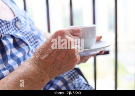 Donna anziana seduta con tazza bianca di caffè in mani rugose. Concetto di mattina fresca, energia e godersi la vita al pensionamento Foto Stock