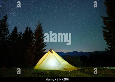 Luminosa tenda turistica illuminata che si illumina sul campeggio in montagne scure sotto il cielo notturno con stelle scintillanti. Concetto di stile di vita attivo Foto Stock