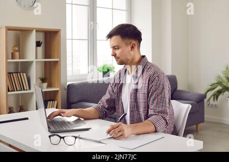 Giovane concentrato che lavora da remoto sul notebook e prende appunti in casa. Foto Stock