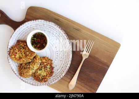 frittelle di zucchine vegetariane in un piatto bianco e salsa asiatica con spezie in una piccola ciotola bianca su un tagliere di legno. Sfondo bianco. Vegetaria Foto Stock