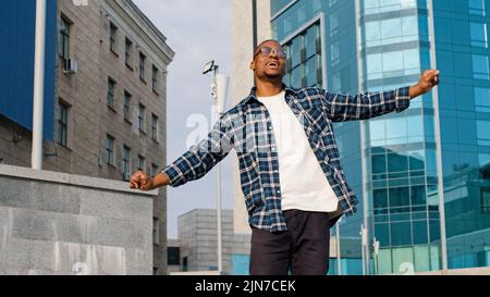 Elegante felice allegro afroamericano ragazzo africano uomo cantare canti canzone godersi la musica ballare all'aperto città edificio urbano sfondo danza avendo Foto Stock