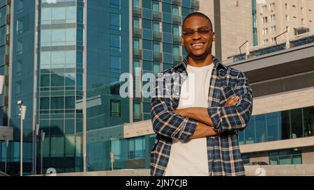 Uomo alla moda in città africano americano ragazzo giovane studente maschio in occhiali da sole indossa camicia plaid in posa con mani incrociate all'aperto guardando macchina fotografica sorridente Foto Stock