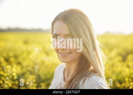 Ritratto di giovane donna con capelli lunghi, campo di fiori gialli e luce solare dietro di lei Foto Stock