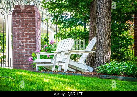 Due sedie Adirondack di legno bianche si siedono da elegante recinzione di ferro con i fiori di Hydrangea in backgound sotto gli alberi Foto Stock