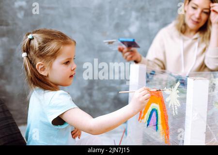 Ritratto di ragazza piccola occupato seduta su lezione d'arte disegno con pennello su film estensibile con l'aiuto di donna tenendo vernici. Foto Stock