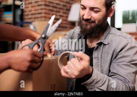 Marito e moglie che tagliano nastro adesivo con forbici per muoversi in appartamento affittato, usando rullo appiccicoso per imballare i mobili per il trasporto di deposito. Giorno commovente per nuovi inizi. Primo piano. Foto Stock