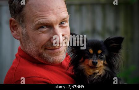 Un uomo maturo che tiene il suo cane chihuahua, sorridendo alla macchina fotografica. Ritratto, testa e spalle, all'aperto. Foto Stock