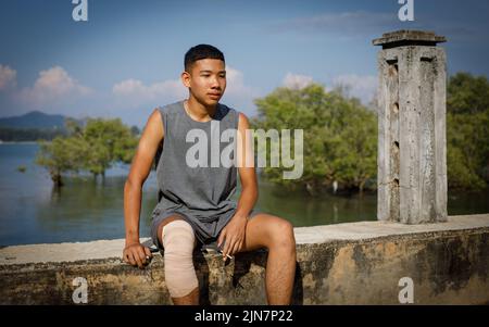 Un adolescente thailandese in una maglietta grigia e una gamba con bendaggi, siede su un muro di mare con una sigaretta in mano con il mare blu e gli alberi verdi dietro Foto Stock