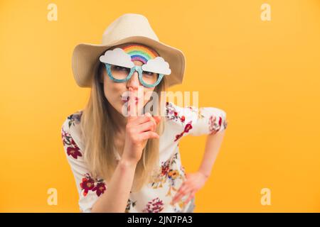 Creativa bella bionda caucasica signora con divertente arcobaleno occhiali che mostrano un segno di silenzio con il suo dito e guardando la fotocamera - sfondo arancione. Foto di alta qualità Foto Stock