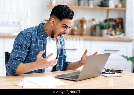 Arrabbiato infastidito giovane uomo, arabo o indiano società lavoratore, freelancer, elegantemente vestito, lavorare da casa, sedersi in cucina a una scrivania, urlare al computer portatile, insoddisfatto del risultato Foto Stock