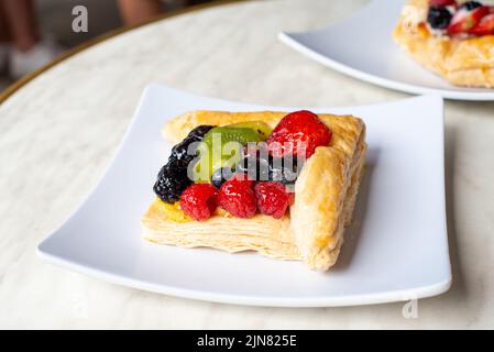 Una sfoglia francese ripiena di frutta quadrata su un piatto quadrato bianco. Il dessert ripieno di crema e crema pasticcera contiene fragole, kiwi e more Foto Stock