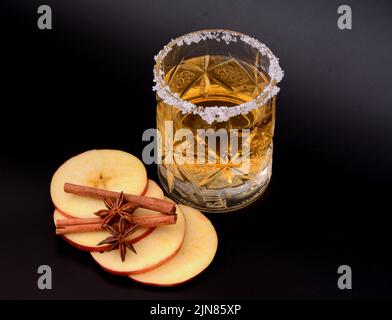 Bicchiere di cristallo con sidro e zucchero intorno ai bordi, accanto a fette di mela, cannella e anice su sfondo nero. Primo piano. Foto Stock