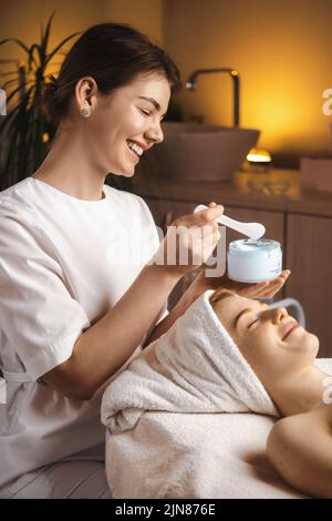 Beautician sorridente che applica la crema al viso di una bella donna nel salone spa. Assistenza sanitaria, trattamenti di bellezza. Donna bellezza viso. Trattamento di bellezza. Foto Stock