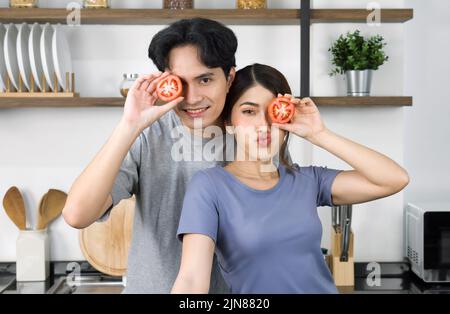 Giovane coppia in vestiolo casual, tenere il pomodoro a fette in posizione occhio. Gli amanti asiatici trascorrono il tempo di mattina insieme in cucina. Foto Stock