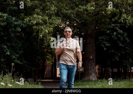 Vecchio uomo brutale in occhiali da sole neri camminando lungo il sentiero nel parco forestale della città in giorno di sole. Stile di vita urbano, tempo libero, vacanza, attività ricreative all'aperto Foto Stock