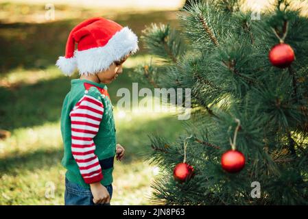 Natale nel mese di luglio. Bambino in attesa di Natale in legno in estate. Ritratto di ragazzo decorazione albero di natale. Vacanze invernali e concetto di persone. Buon Natale e buone feste Foto Stock