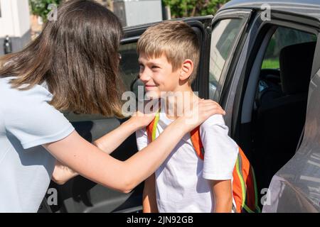 La mamma abbraccia e mette il bambino con una borsa da scuola in auto da portare a scuola. Concetto di ritorno a scuola. Foto Stock