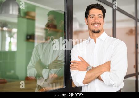 Sorridente latino alto leader esperto in abbigliamento formale guarda da parte, fiducioso ambizioso barbuto uomo d'affari ispanico si trova in ufficio con le braccia piegate, proprietario startup Foto Stock