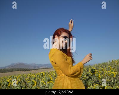 Vista laterale della femmina positiva in occhiali da sole che ballano con braccia sollevate mentre si trova in piantagione con girasoli gialli in campagna Foto Stock