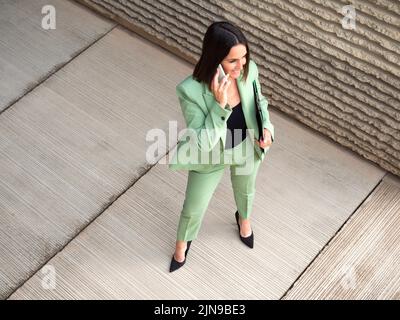 Da sopra il corpo pieno di imprenditrice femminile positiva in elegante vestito con cartella che parla sullo smartphone mentre si trova in piedi sulla strada Foto Stock