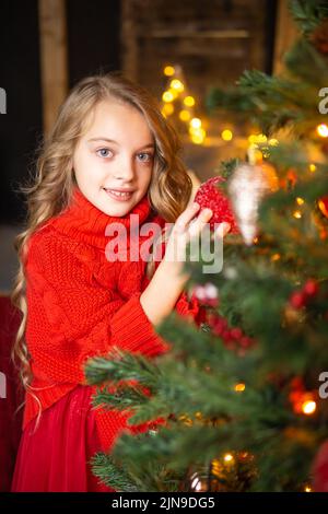 Una bambina bionda in un abito festivo accanto ad un albero di Natale decorato con ghirlande, palle di vetro e giocattoli di Natale. Il concetto di holid invernale Foto Stock