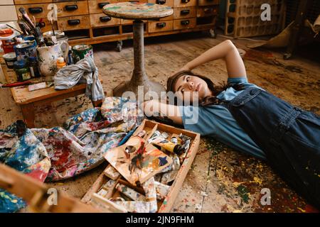 Giovane donna artista sdraiato sul pavimento mentre lavora sul pavimento nel suo studio interno Foto Stock