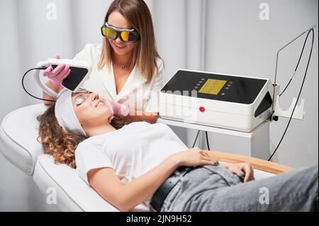 Cosmetologa femminile che utilizza un dispositivo di scansione laser a diodi durante la procedura di rifacing skincare. Donna sorridente paziente in occhiali protettivi che riceve un trattamento laser del viso in salone di bellezza. Foto Stock