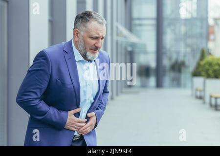 Uomo d'affari anziano grigio-haired al di fuori della costruzione dell'ufficio, uomo nel vestito di affari malato ha stomachache. Foto Stock