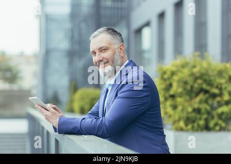 Ritratto di successo capo uomo d'affari senior dai capelli grigi, uomo fuori ufficio utilizzando tablet computer sorridendo e guardando la fotocamera, investitore in tuta d'affari, banchiere felice Foto Stock