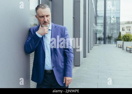 Uomo grigio senior capelli fuori ufficio edificio, in piedi e pensare, confusa e triste businessman boss in tuta d'affari, su decisione Foto Stock