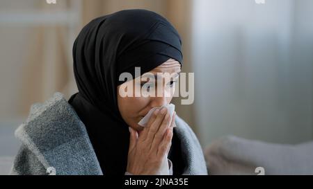 Primo piano malato sconvolto giovane ragazza in hijab starnutire naso con carta tovagliolo triste non sano musulmana che beve tè caldo che soffre di freddo stagionale Foto Stock