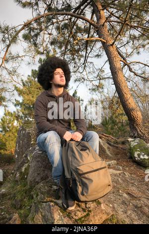 uomo con capelli afro seduto su una roccia nella foresta guardando l'orizzonte Foto Stock