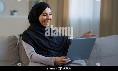 Amichevole ragazza musulmana araba in hijab sedersi sul divano accogliente parlare videochiamata con amico saluto al computer webcam prendere intervista distanza virtuale Foto Stock