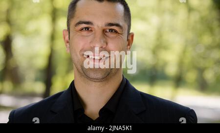 Outdoors ritratto di uomo adulto caucasico ragazzo felice maschio faccia riuscito uomo d'affari millenario indossa vestito nero girando testa guardando la macchina fotografica sorridente Foto Stock