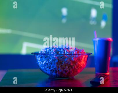 Telecomando TV, popcorn in una ciotola e un drink in un bicchiere sullo sfondo di una partita di calcio in TV. Illuminazione al neon. Riposo confortevole a casa guardare Foto Stock