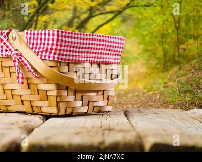 Un cestino da picnic di paglia su un semplice tavolo di legno sullo sfondo di una meravigliosa natura autunnale. Il sole d'autunno illumina le foglie dorate sul Foto Stock