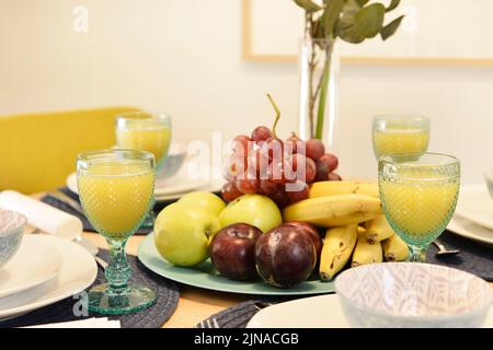 Centrotavola con vassoio di frutta fresca, vaso decorativo, grappolo d'uva, tante banane, mele dorate e bicchieri di succo d'arancia Foto Stock