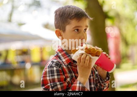 Primo piano di un piccolo simpatico ragazzo caucasico che mangia deliziosi waffle in strada. Bambino con fast food. Foto Stock