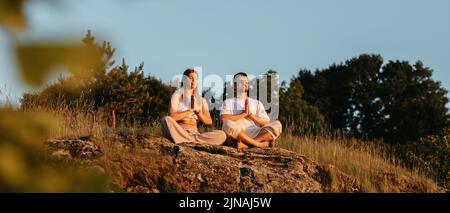 Uomo e donna meditando all'aperto mentre si siede sulla roccia, giovane coppia adulta pratica lo yoga al tramonto Foto Stock