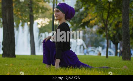Musulmana ragazza islamica in hijab allenatore guru yoga master donna fare esercizi di stretching nel parco su erba verde fare sport si siede in bella acrobatica Foto Stock