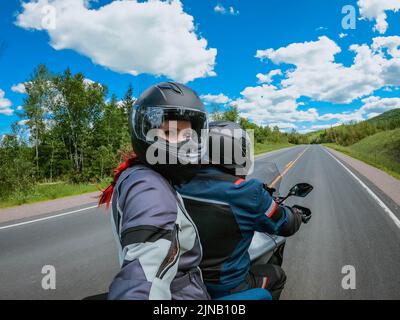 La giovane donna del passeggero del motociclo in un casco fa selfie sulla macchina fotografica di azione mentre guida sul retro del motociclo su una strada di paesaggio vuota. Foto Stock