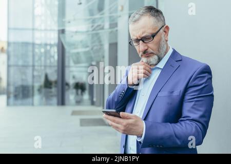 Pensiero senior grigio-haired investitore leggere le notizie sul telefono, uomo d'affari in piedi fuori ufficio edificio e tenendo smartphone, uomo premuroso e serio in occhiali e tuta aziendale Foto Stock