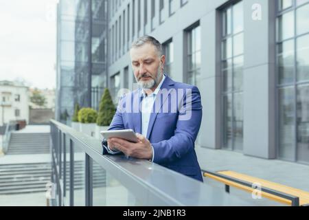 Riuscito e serio uomo grigio haired senior fuori ufficio edificio utilizzando tablet computer, imprenditore investitore leggere notizie dal tablet Foto Stock