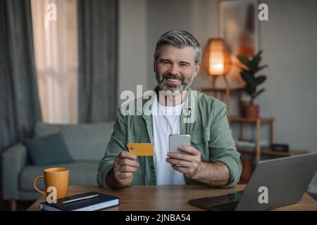 Acquista e paga a casa. Sorridente uomo europeo in pensione con barba usa smartphone, computer e carta di credito Foto Stock